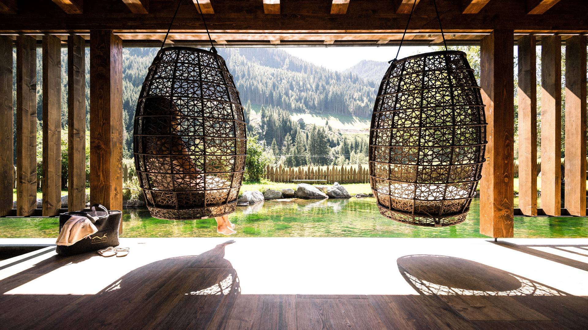 Peace in your own rhythm Two large hanging chairs made of woven rattan overlooking a natural pond and forested mountains in the background.